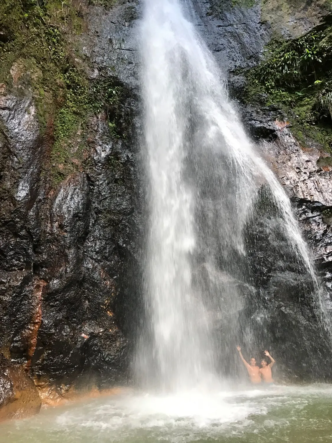 Two of the group waving under a tall waterfall in Dominica